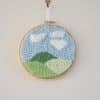 Décoration murale pour enfant. Cadre réalisé à la broderie punchneedle représentant un paysage : 3 collines. Chaque colline a un ton de vert différent. Le ciel est bleu. Il y a des nuages blancs.