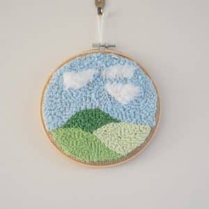 Décoration murale pour enfant. Cadre réalisé à la broderie punchneedle représentant un paysage : 3 collines. Chaque colline a un ton de vert différent. Le ciel est bleu. Il y a des nuages blancs.