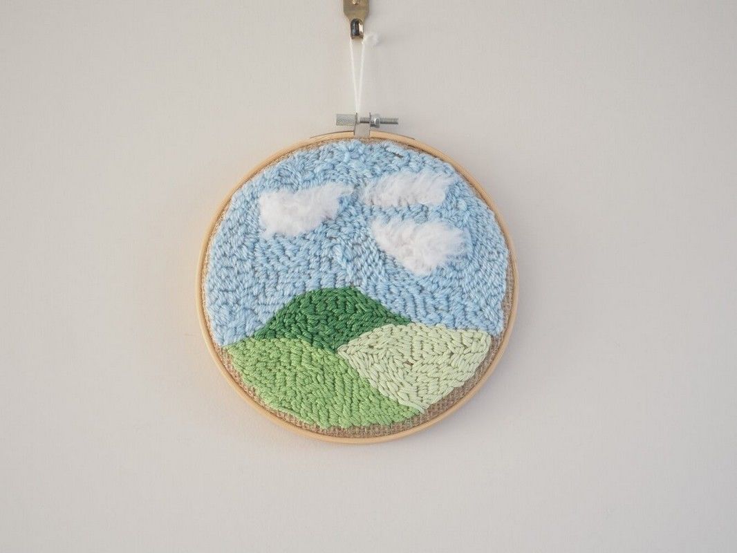 Décoration murale pour enfant. Cadre réalisé à la broderie punchneedle représentant un paysage : 3 collines. Chaque colline a un ton de vert différent. Le ciel est bleu. Il y a des nuages blancs.