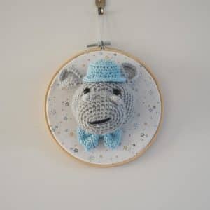 Décoration murale de type trophée au crochet. Hippopotame avec noeud papillon et chapeau bleu clair. Trophée monté sur un tambour. Fond du tambour en tissus avec des étoiles de couleurs moutarde, bleu clair, bleu marine, moutarde.