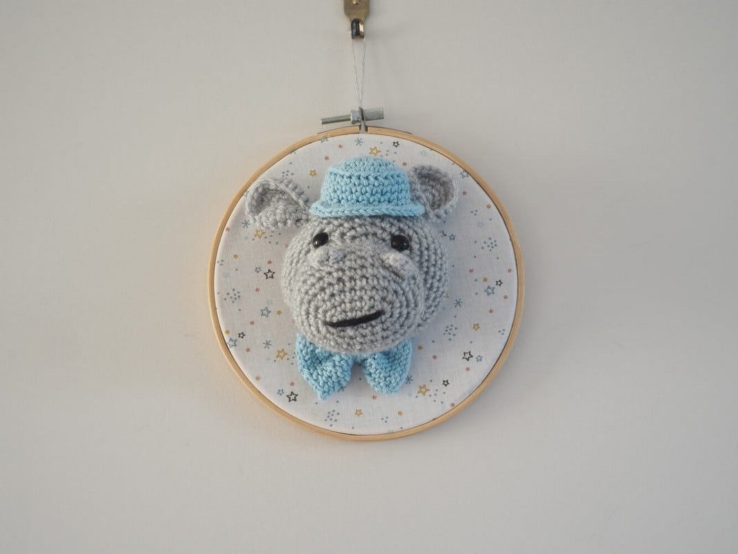 Décoration murale de type trophée au crochet. Hippopotame avec noeud papillon et chapeau bleu clair. Trophée monté sur un tambour. Fond du tambour en tissus avec des étoiles de couleurs moutarde, bleu clair, bleu marine, moutarde.