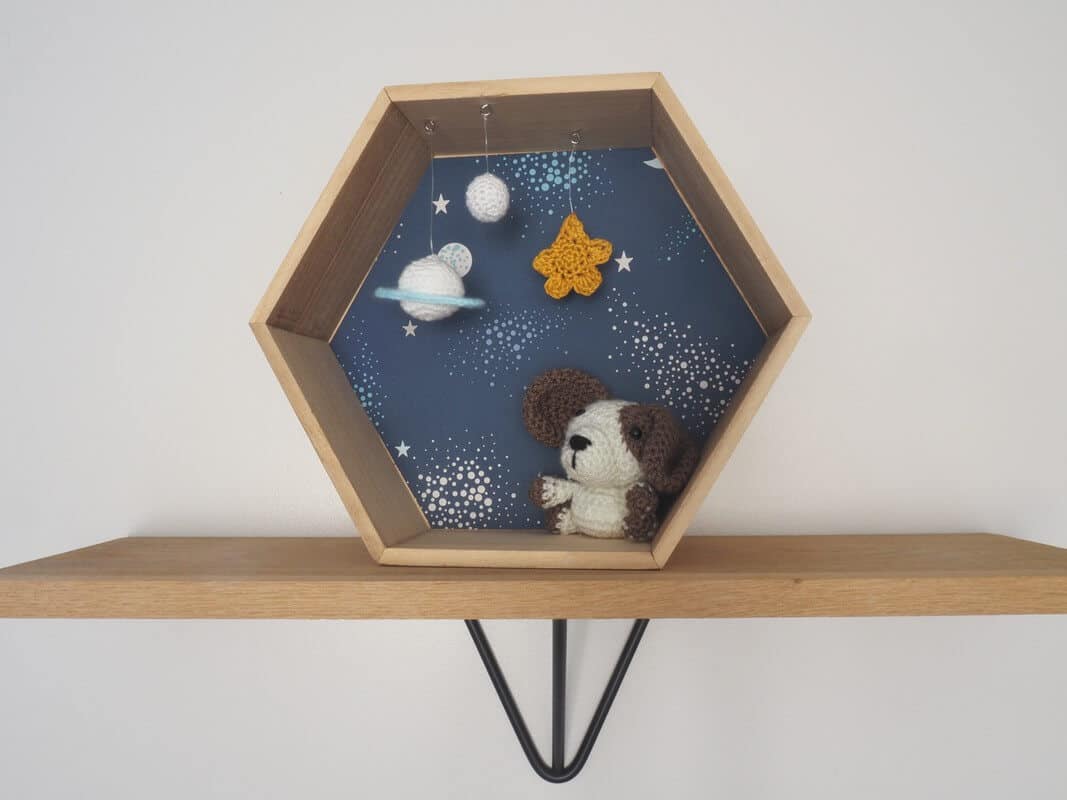 Décoration enfant. Petit chien réalisé au crochet dans cadre hexagonal. Le fond du cadre est tapissé d'un papier peint représentant des étoiles et des planètes. Le personnage au crochet est fixé au cadre. Suspendu au cadre 2 planètes et une étoile réalisés au crochet.