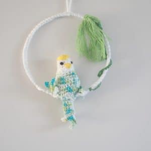 Cadre mural. Ce cadre est composé d’un cadre métallique agrémenté de feuillages verts (laine coton) et d’une perruche dodue réalisée au crochet. Le corps, les ailes et la queue de la perruche sont multi couleurs : bleu, vert, blanc. La tête est blanche avec un bec jaune et un plumage jaune sur la tête.