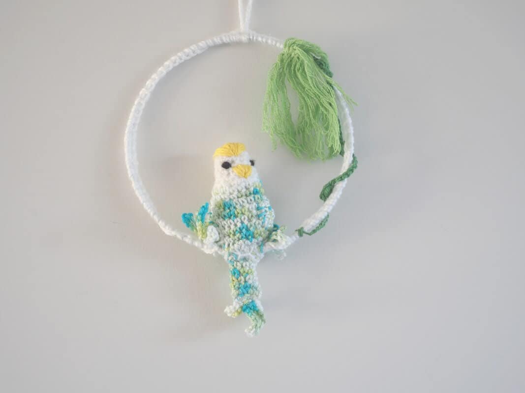 Cadre mural. Ce cadre est composé d’un cadre métallique agrémenté de feuillages verts (laine coton) et d’une perruche dodue réalisée au crochet. Le corps, les ailes et la queue de la perruche sont multi couleurs : bleu, vert, blanc. La tête est blanche avec un bec jaune et un plumage jaune sur la tête.