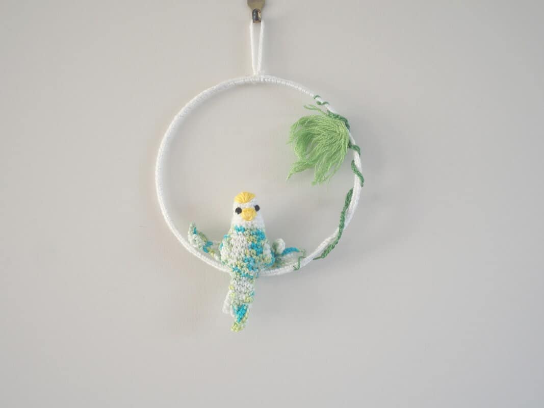 Cadre mural. Ce cadre est composé d’un cadre métallique agrémenté de feuillages verts (laine coton) et d’une perruche réalisée au crochet. Le corps, les ailes et la queue de la perruche sont multi couleurs : bleu, vert, blanc. La t^te est blanche avec un bec jaune et un plumage jaune sur la tête.