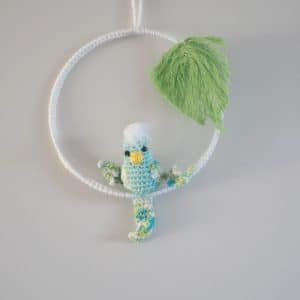 Cadre mural. Ce cadre est composé d’un cadre métallique agrémenté d'un feuillage (laine coton) et d’une perruche réalisée au crochet. La perruche est de couler vert d'eau pour le corps. Les ailes et la queues sont bleues, vertes et blanches. Le bec est jaune et le plumage sur la tête blanche.