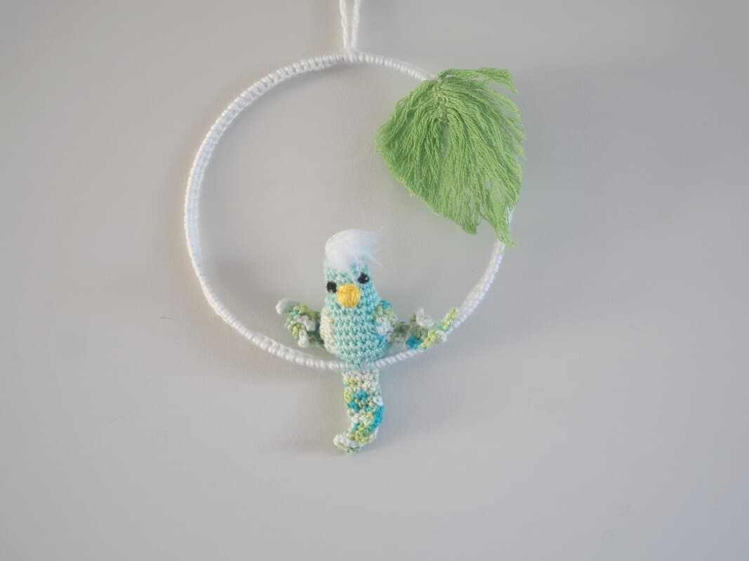 Cadre mural. Ce cadre est composé d’un cadre métallique agrémenté d'un feuillage (laine coton) et d’une perruche réalisée au crochet. La perruche est de couler vert d'eau pour le corps. Les ailes et la queues sont bleues, vertes et blanches. Le bec est jaune et le plumage sur la tête blanche.