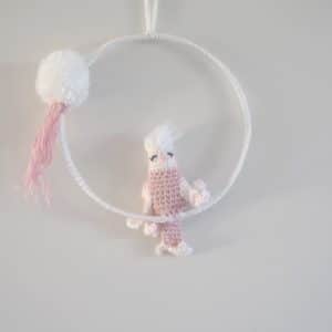 Cadre mural. Ce cadre est composé d’un cadre métallique agrémenté d'un pompon blanc, d'un feuillage rose et d’une perruche réalisée au crochet. La perruche est de couleur rose pour le corps. Les ailes, la queue et la tête sont blanches. Le bec est rose et le plumage sur la tête blanc.