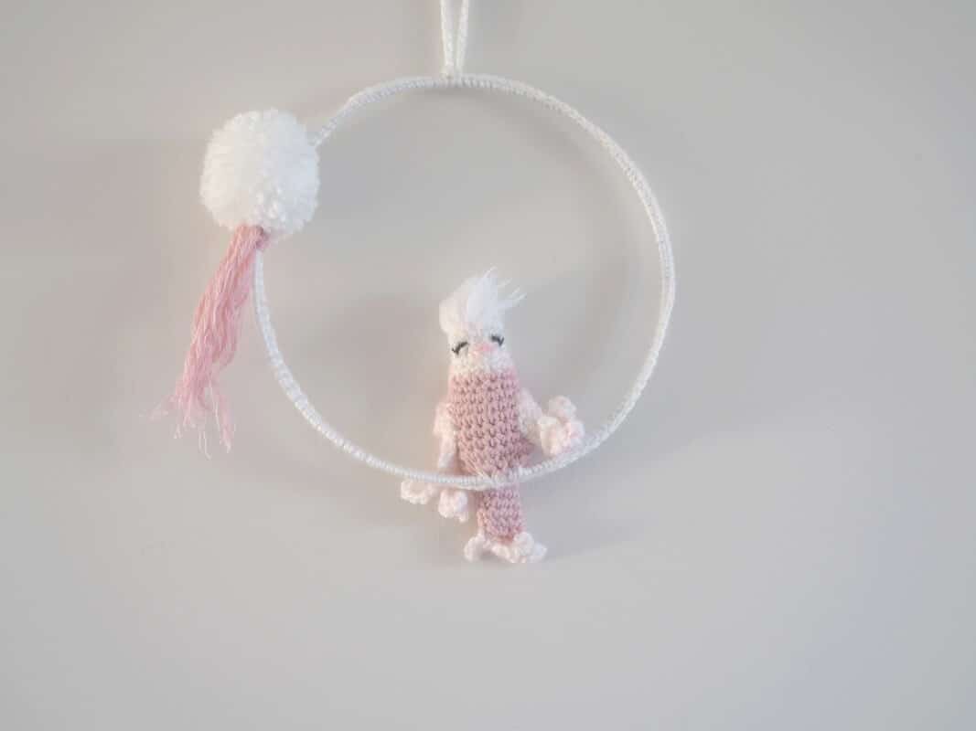 Cadre mural. Ce cadre est composé d’un cadre métallique agrémenté d'un pompon blanc, d'un feuillage rose et d’une perruche réalisée au crochet. La perruche est de couleur rose pour le corps. Les ailes, la queue et la tête sont blanches. Le bec est rose et le plumage sur la tête blanc.