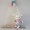 Décoration murale. Lapine blanche en crochet vêtue d’une robe en tissu liberty ( fleurs roses et bleues) et de chaussures et écharpe rose. Le personnage est fixé sur une balancelle en bois. Pompons bleu, blanc, rose fixés au cordage.