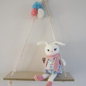 Décoration murale. Lapine blanche en crochet vêtue d’une robe en tissu liberty ( fleurs roses et bleues) et de chaussures et écharpe rose. Le personnage est fixé sur une balancelle en bois. Pompons bleu, blanc, rose fixés au cordage.