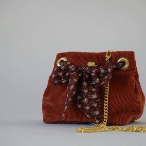 Sac seau en velours orange brûlé / rouille porté croisé par une chaine dorée. Fermeture par un bandeau en tissu marron, petites fleurs blanches.