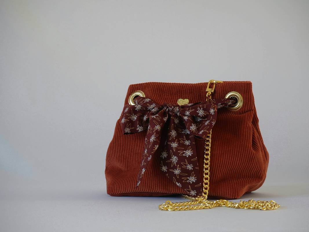 Sac seau en velours orange brûlé / rouille porté croisé par une chaine dorée. Fermeture par un bandeau en tissu marron, petites fleurs blanches.