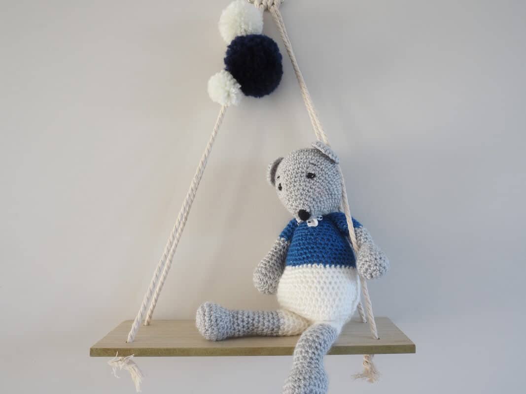 Souris grise, blanc et bleue au crochet fixée sur une balancelle en bois. Pompons blancs et bleu marine fixés au cordage.