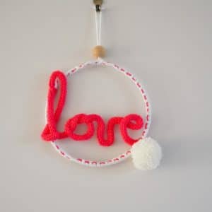 Décoration murale. Mot love en tricotin, de couleur rose. Pompon blanc. Le mot love est fixé à un cercle métallique.