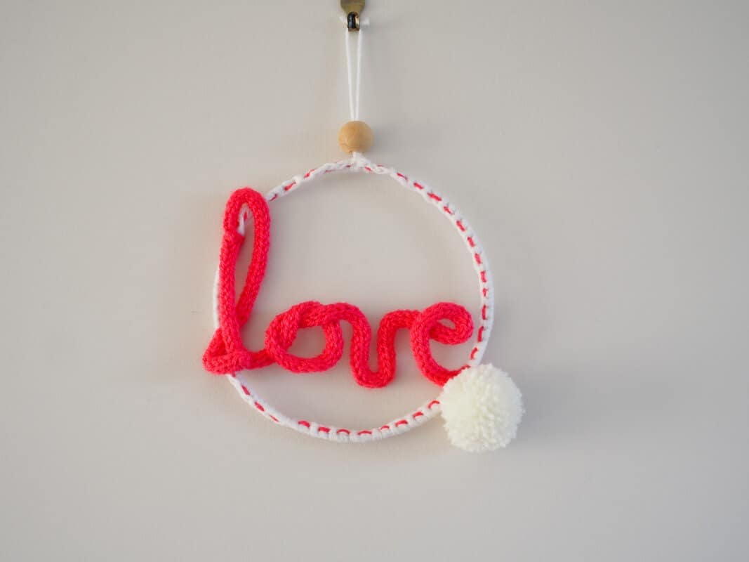 Décoration murale. Mot love en tricotin, de couleur rose. Pompon blanc. Le mot love est fixé à un cercle métallique.