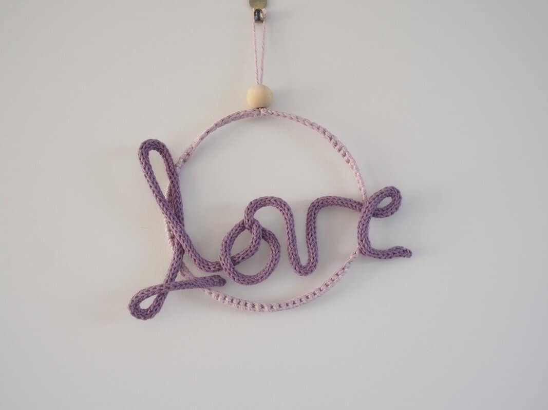 Décoration murale. Mot love en tricotin, de couleur violette. Le mot love est fixé à un cercle métallique.