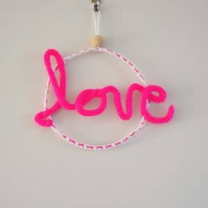 Décoration murale. Mot love en tricotin, de couleur rose fluo. Le mot love est fixé à un cercle métallique.