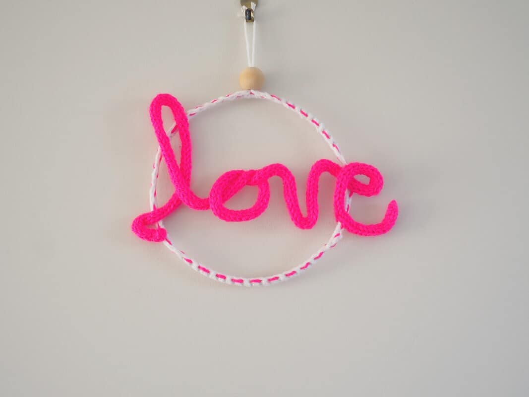 Décoration murale. Mot love en tricotin, de couleur rose fluo. Le mot love est fixé à un cercle métallique.