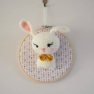 Décoration murale de type trophée au crochet. Lapine avec noeud papillon moutarde. Trophée monté sur un tambour. Fond du tambour en tissus avec des points de couleur rose, moutarde.