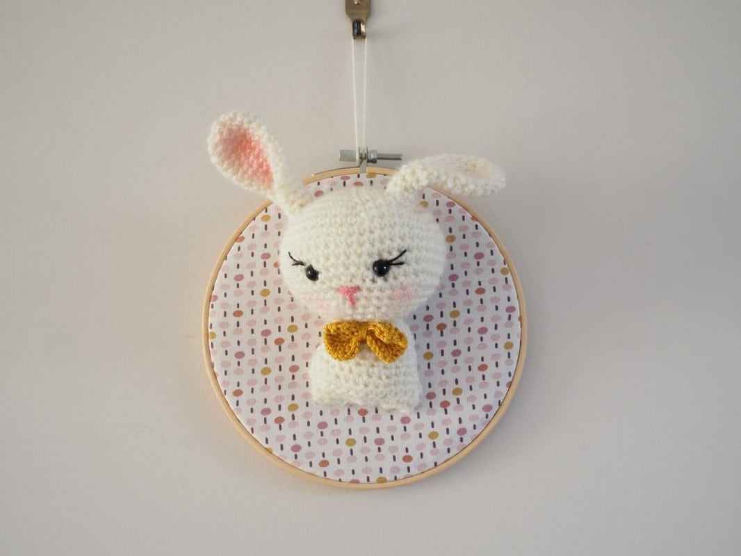 Décoration murale de type trophée au crochet. Lapine avec noeud papillon moutarde. Trophée monté sur un tambour. Fond du tambour en tissus avec des points de couleur rose, moutarde.