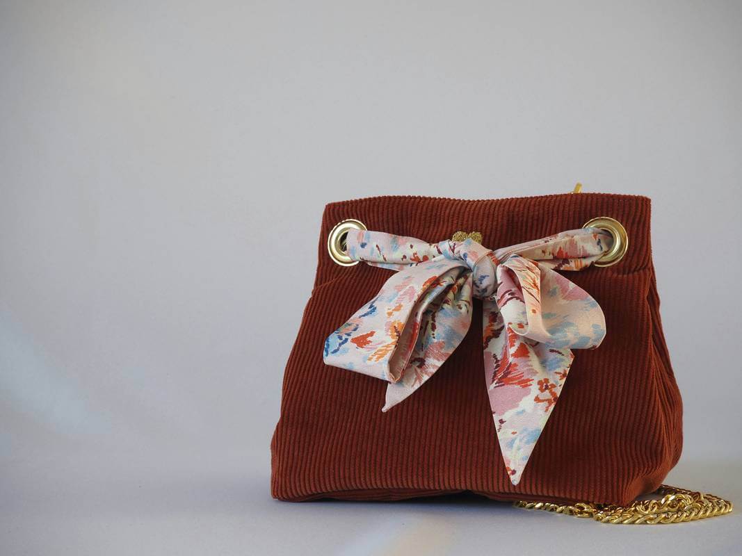 Sac seau en velours orange brûlé / rouille porté croisé par une chaine dorée. Fermeture par un bandeau en tissu multicolore rosé orangé.