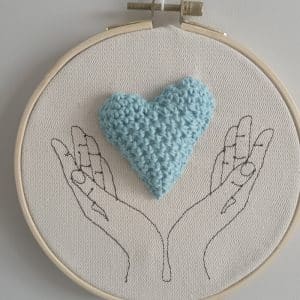 Cadre représentant des mains (broderie). Entre les mains se trouve un coeur réalisé au crochet.