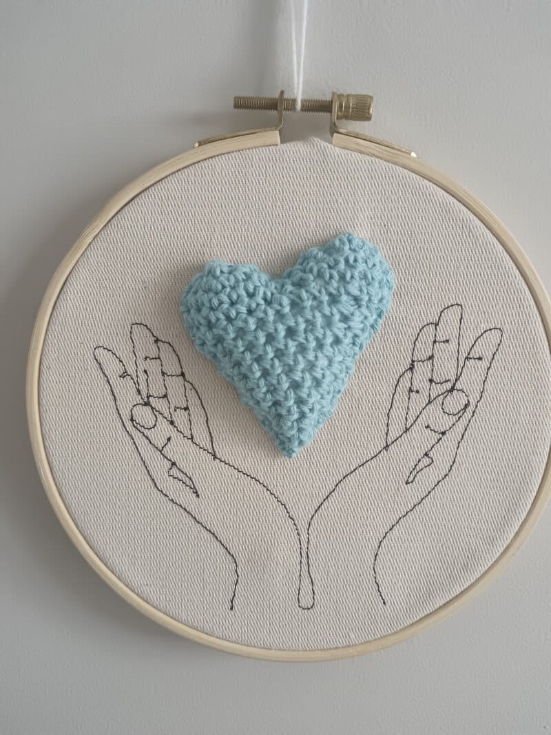 Cadre représentant des mains (broderie). Entre les mains se trouve un coeur réalisé au crochet.