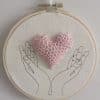 Cadre représentant des mains (broderie). Entre les mains se trouve un coeur réalisé au crochet.