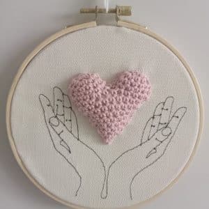 Cadre représentant des mains (broderie). Entre les mains se trouve un coeur réalisé au crochet.