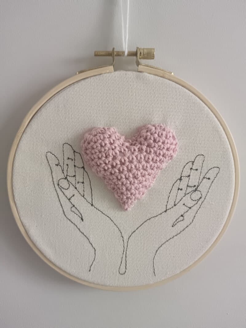 Cadre représentant des mains (broderie). Entre les mains se trouve un coeur réalisé au crochet.