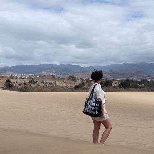 Sac cabas noir à motif tropical porté avec une tenue estivale beige / écrue, dans un décor de dune.