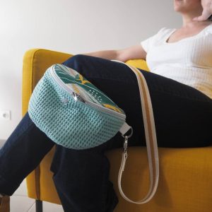 Sac banane Lagune en coton. Corps du sac en nid d'abeille couleur sauge et rabat tropique multicouleur. Portée avec une bandoulière moutarde et liserés argentés.