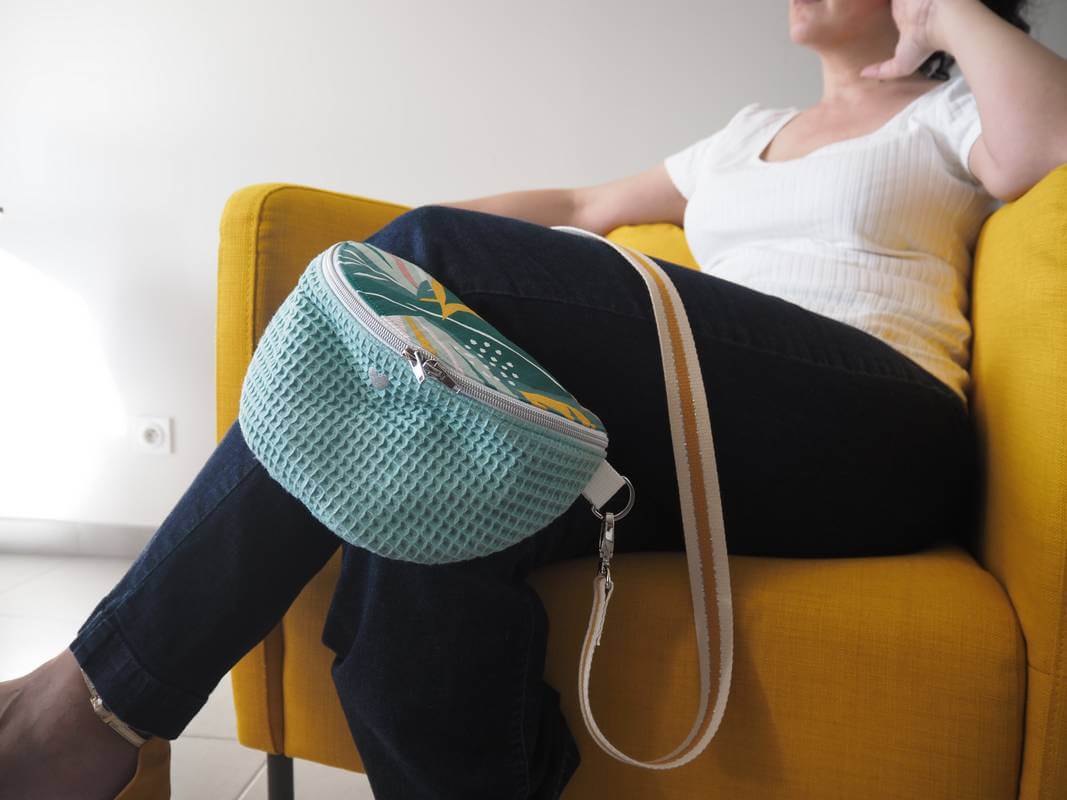 Sac banane Lagune en coton. Corps du sac en nid d'abeille couleur sauge et rabat tropique multicouleur. Portée avec une bandoulière moutarde et liserés argentés.