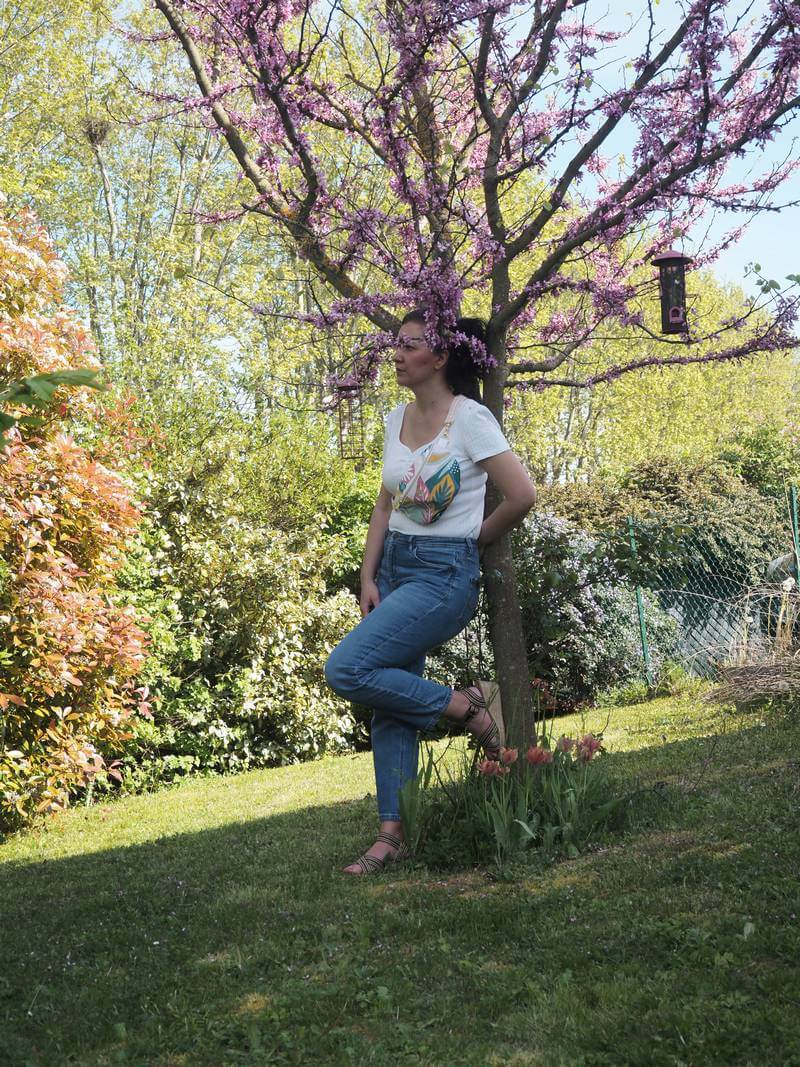 Sac banane "Tropique" avec des motifs tropicaux et multi color : rose, vert, sauge, moutarde ... Ce sac banane est porté en cross body sur un haut blanc, un pantalon en jean et avec des sandales compensées.
