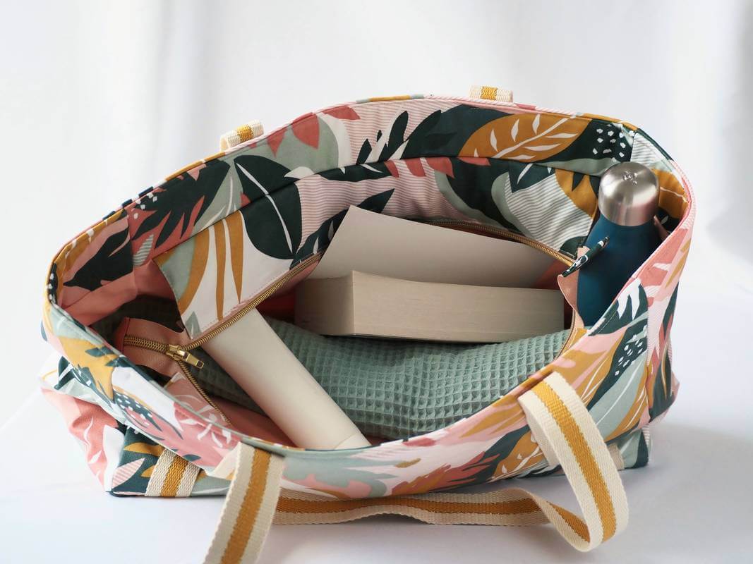Sac cabas en coton enduit, de cmotif tropical multicolor (rose, vert, moutarde,...) , avec fermeture par zip. Présenté avec des affaires de plage.